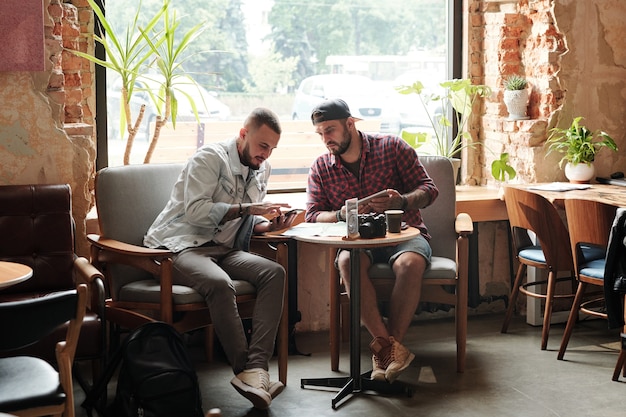 Amigos varones jóvenes sentados a la mesa en la cafetería loft y hablando sobre un nuevo viaje mientras usan dispositivos modernos