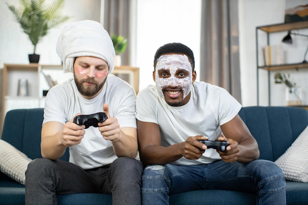 Amigos varones con máscaras faciales y parches para los ojos sentados en el sofá y jugando videojuegos