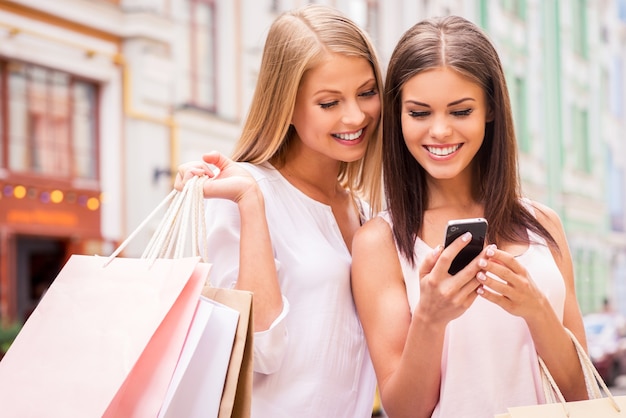 Amigos viciados em compras. Duas mulheres atraentes segurando sacolas de compras e olhando para o celular juntas enquanto ficam do lado de fora