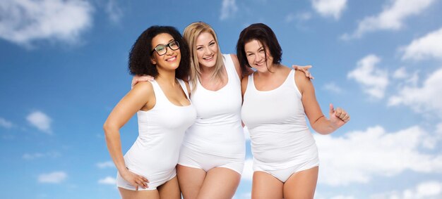 amistad, belleza, cuerpo positivo y concepto de personas - grupo de mujeres felices de talla grande en ropa interior blanca sobre cielo azul y fondo de nubes