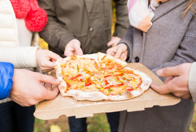 Amistad, comida chatarra y concepto de personas: cerca de las manos de los amigos comiendo pizza de una caja de cartón al aire libre