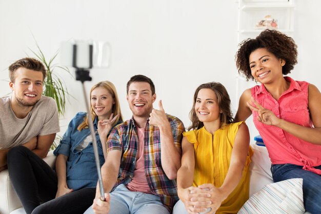 amistad y concepto de la gente - grupo de amigos felices tomando fotos con smartphone y selfie stick en casa