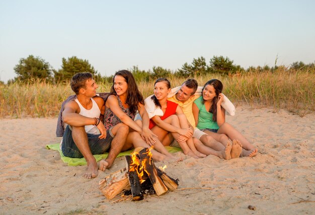 amistad, felicidad, vacaciones de verano, vacaciones y concepto de la gente - grupo de amigos sonrientes sentados cerca del fuego en la playa