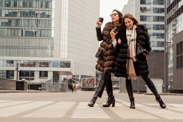 Amistad femenina, relaciones. Dos chicas beben café en la calle cerca de edificios de oficinas de vidrio, una corporación, un banco. El final de la jornada laboral del gerente. Café a la carrera.