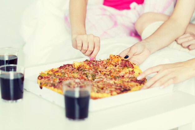 Foto amistad, gente, fiesta de pijamas y concepto de comida chatarra - cerca de amigos o adolescentes comiendo pizza en casa
