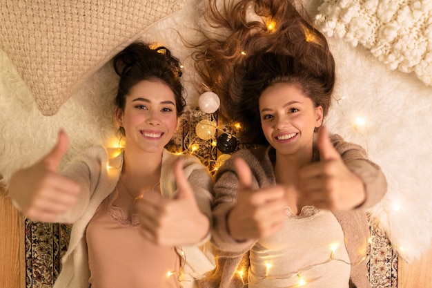 Foto amistad hygge y concepto de fiesta de pijama amigas felices o niñas adolescentes acostadas en el suelo y mostrando los pulgares hacia arriba en casa