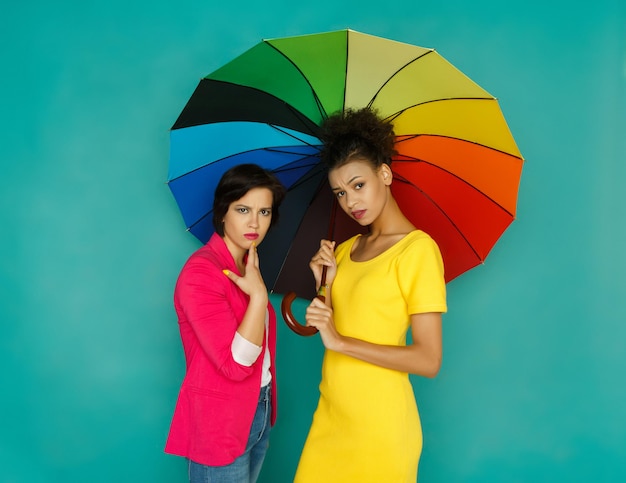 Amistad multiétnica. Chicas negras y caucásicas preocupadas en ropa casual colorida chismeando bajo el paraguas de colores del arco iris en el fondo azul del estudio con espacio de copia