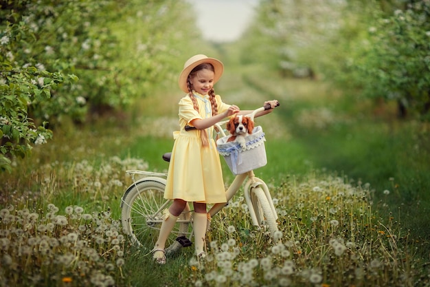 La amistad de los niños y las mascotas, una linda chica y un pequeño cachorro, montan en bicicleta en primavera en un jardín floreciente