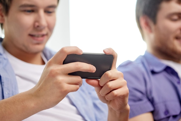 Amistad, tecnología y concepto de personas: cerca de amigos varones sonrientes felices con teléfonos inteligentes enviando mensajes de texto o jugando juegos en casa