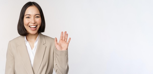 Amistosa mujer de negocios asiática dama de la oficina saludando con la mano y diciendo hola hola gesto de pie sobre fondo blanco.