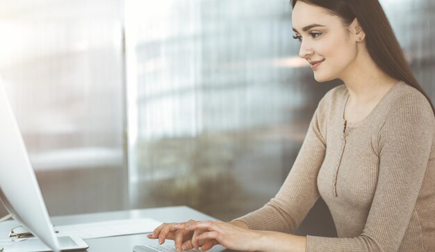 Una amistosa mujer de negocios vestida de forma informal está trabajando en su computadora, mientras está sentada en el escritorio en una oficina soleada. Concepto de éxito empresarial