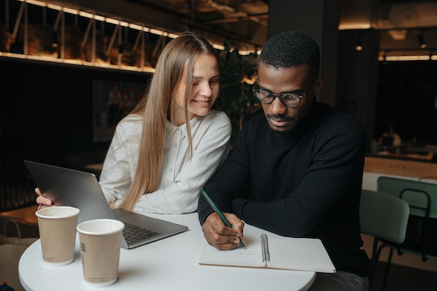 Amistosos colegas interraciales del sexo opuesto en el café trabajando juntos en una computadora portátil, mientras escriben notas.