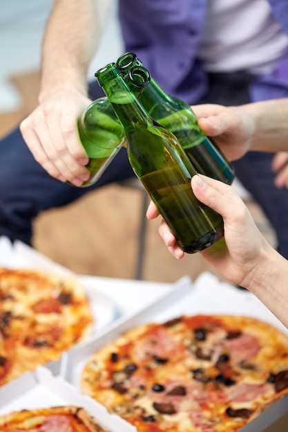 amizade, álcool, pessoas, conceito de celebração e feriados - close-up de mãos masculinas tilintando garrafas de cerveja e comendo pizza em casa
