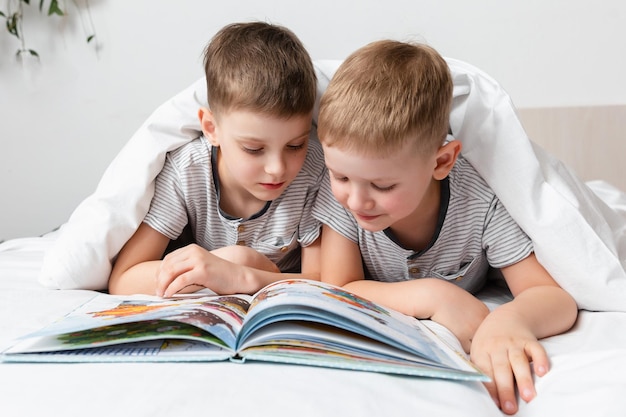 Amizade de irmãos Duas crianças felizes e fofas se divertem quando lêem um livro em uma cama debaixo de um cobertor Educação em casa Atividade interna