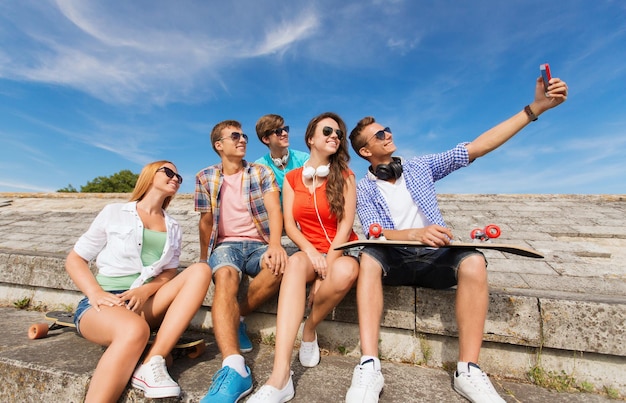 amizade, lazer, verão, tecnologia e conceito de pessoas - grupo de amigos sorridentes com skate e smartphone fazendo selfie ao ar livre