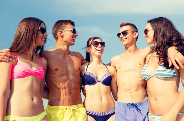 amizade, mar, férias de verão, feriados e conceito de pessoas - grupo de amigos sorridentes vestindo roupas de banho e óculos de sol conversando e rindo na praia