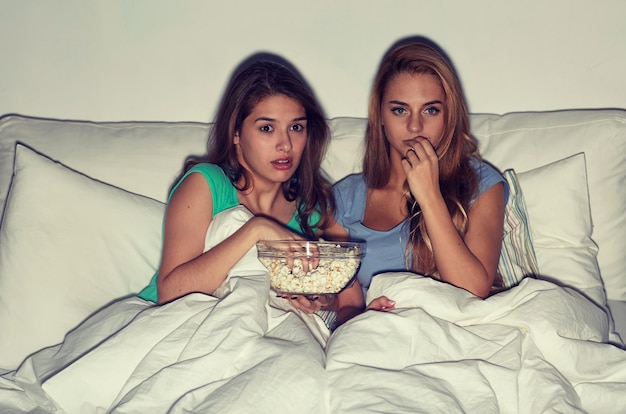 amizade, pessoas, festa do pijama, entretenimento e conceito de junk food - amigos assustados ou adolescentes comendo pipoca e assistindo filme de terror na tv em casa
