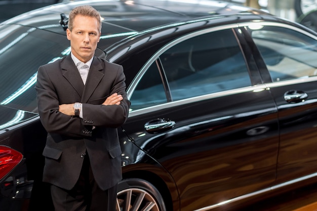 Amo los autos de lujo. Vista superior del hombre de pelo gris confiado en ropa formal apoyado en el coche y mirando a la cámara