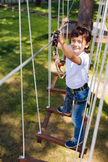 Amo las aventuras. La vista superior de un alegre niño preadolescente escalando en un parque de cuerdas y sonriendo a la cámara mientras se mueve por senderos