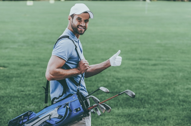 Amo jogar golfe! Retrovisor de um jovem jogador de golfe feliz carregando uma sacola de golfe com motoristas e olhando por cima do ombro enquanto está de pé no campo de golfe