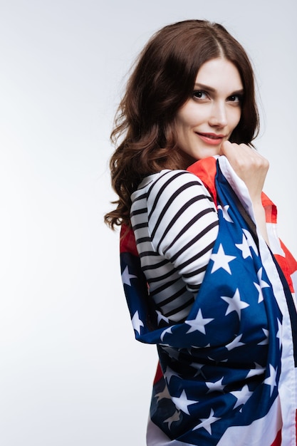 Amo a mi nación. Bonita mujer joven de pelo castaño posando envuelta en una bandera americana