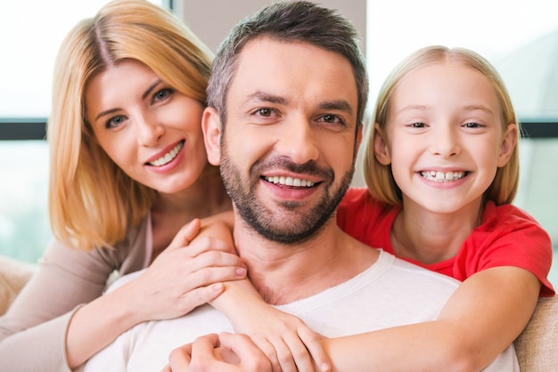 Foto ¡amo a mis chicas! familia feliz de tres uniéndose entre sí y sonriendo mientras está sentado en el interior