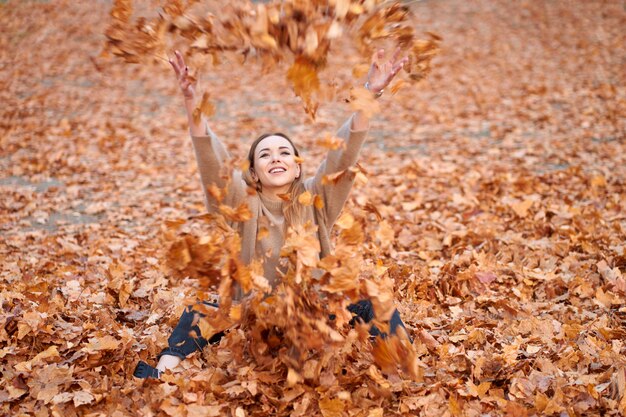 Amo el otoño. Chica atractiva alegre con suéter, jeans negros y botas de cuero negro sentado y lanzando hojas de otoño en el parque de otoño.