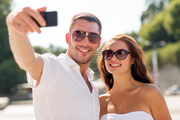 amor, boda, verano, citas y concepto de personas - pareja sonriente con gafas de sol abrazándose y haciendo selfie con smartphone en el parque