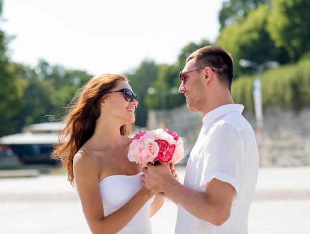 amor, casamento, verão, namoro e conceito de pessoas - casal sorridente usando óculos escuros com ramo de flores olhando um ao outro na cidade