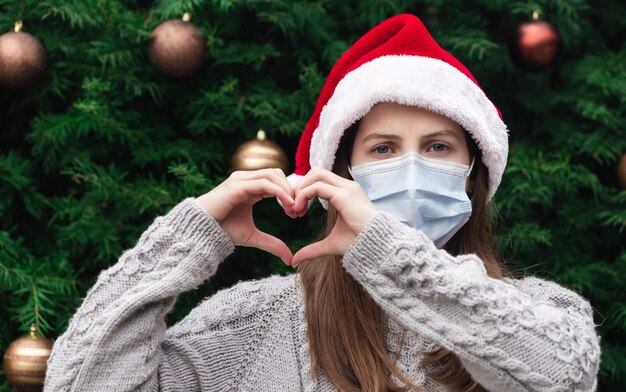 Amor de corazón en forma de mano. Close up Retrato de mujer con un sombrero de santa claus y una máscara médica con emoción. En el contexto de un árbol de Navidad. Pandemia de coronavirus