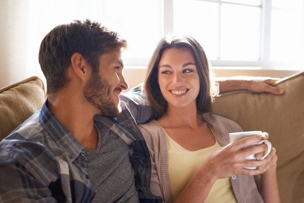 Amor de café e casal sorrindo um para o outro ou em um sofá na sala de estar ou se unindo e conversando Homem mulher e se comunicando ou afetuoso com bebidas quentes ou parceiros felizes em casa