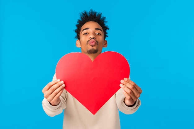 Amor, día de San Valentín y concepto de relación. Chico afroamericano novio lindo con corte de pelo afro, dale a la tarjeta de San Valentín un corazón grande y labios plegables para besar, confesar, buscar alma gemela, pared azul