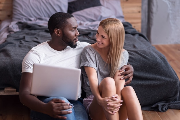 Amor e cuidado. doce casal internacional jovem sentado no quarto juntos, usando o laptop e sorrindo.