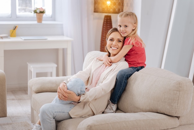 Amor familiar. Linda chica sonriente agradable sentada en el sofá y abrazando a su madre mientras está en casa junto con ella