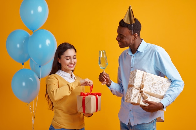 Amor feliz pareja en gorras con vasos de bebidas y cajas de regalo, fondo amarillo. Bonita fiesta familiar, evento o celebración de cumpleaños, decoración de globos.