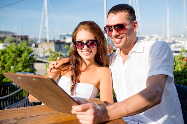 Amor, namoro, pessoas e conceito de comida - casal sorridente usando óculos escuros olhando o menu no terraço do café