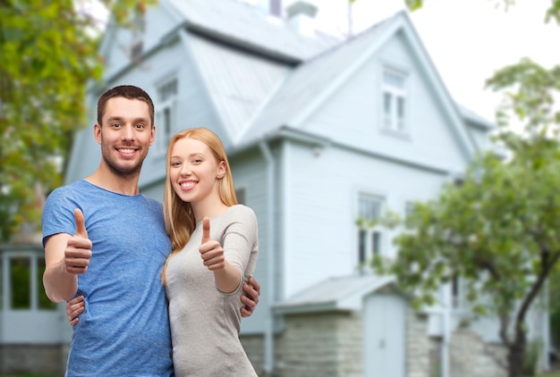 amor, pessoas, imóveis, casa e conceito de família - casal sorridente abraçando e mostrando os polegares sobre o fundo da casa