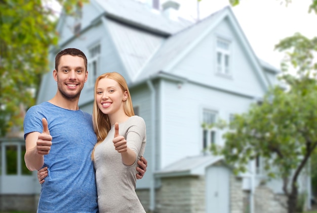 Foto amor, pessoas, imóveis, casa e conceito de família - casal sorridente abraçando e mostrando os polegares sobre o fundo da casa