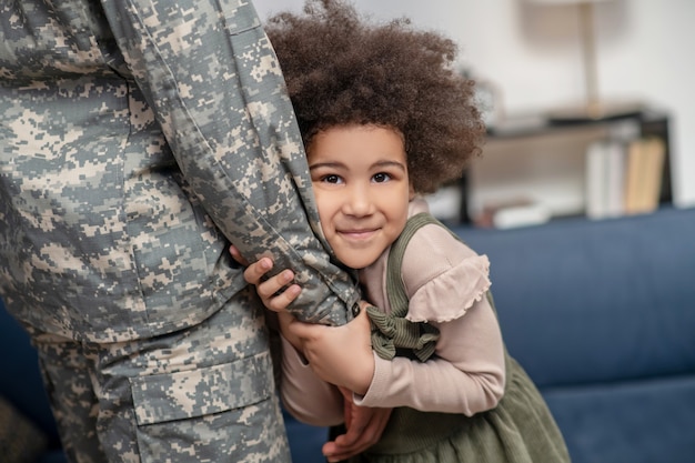 Amor. Poco rizado linda niña afroamericana abrazando la mano de los papás en uniforme militar de pie sonriendo soñando