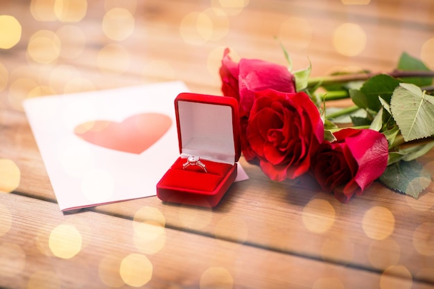 amor, proposta, dia dos namorados e conceito de férias - close-up da caixa de presente com anel de noivado de diamante, rosas vermelhas e cartão em madeira