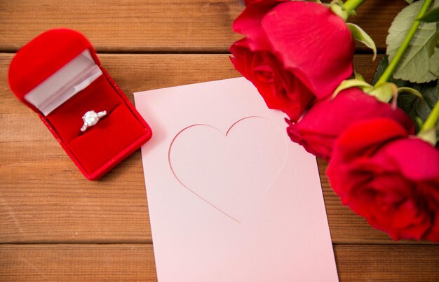 amor, propuesta, día de san valentín y concepto de vacaciones - cierre de caja de regalo con anillo de compromiso de diamantes, rosas rojas y tarjeta de felicitación en madera