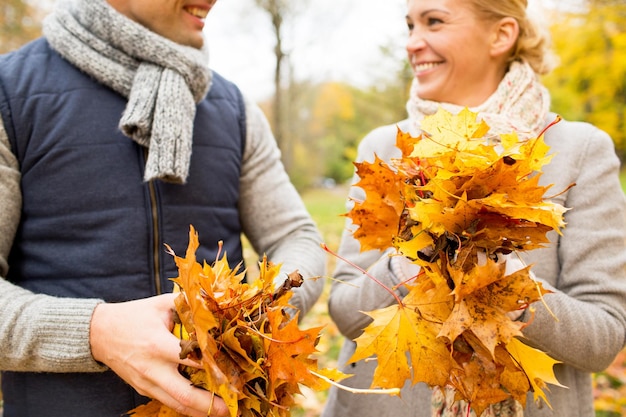 El amor, la relación, la familia, la temporada y el concepto de la gente: cerca de una feliz pareja sonriente con un montón de hojas de arce divirtiéndose en el parque de otoño