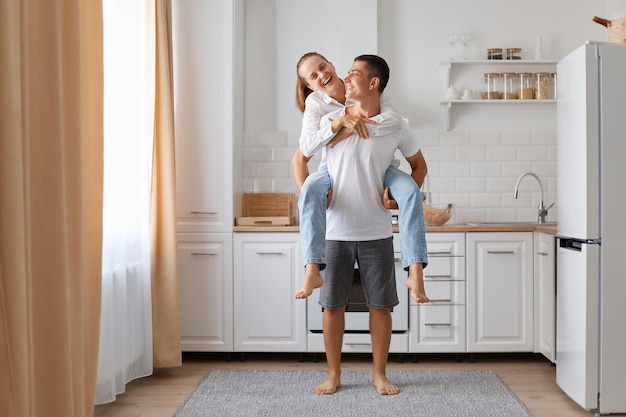 Amor y relación familiar Sonriente joven pareja caucásica con camisas blancas divirtiéndose, el marido sostiene a la esposa de espaldas en el acogedor interior de la cocina Hombres y mujeres disfrutan del tiempo libre juntos en casa