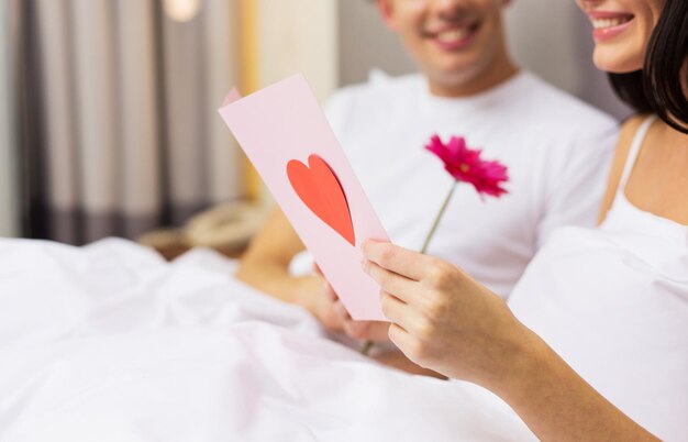 amor, relaciones, vacaciones, día de san valentín y concepto de la gente - cerca de una pareja feliz en la cama con postal y flor