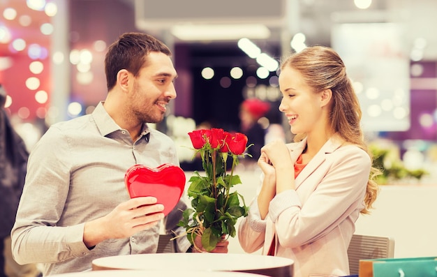 Amor, romance, dia dos namorados, casal e conceito de pessoas - jovem feliz com flores vermelhas dando presente para mulher sorridente no café no shopping