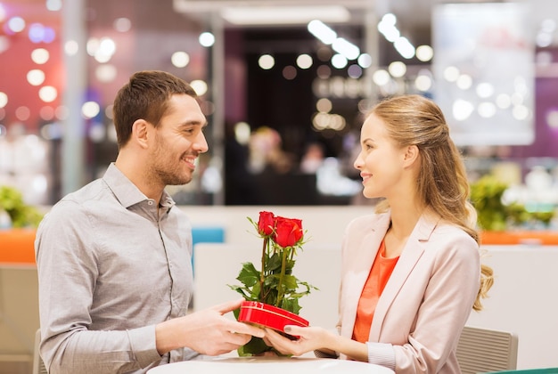 amor, romance, dia dos namorados, casal e conceito de pessoas - jovem feliz com flores vermelhas dando presente para mulher sorridente no café no shopping