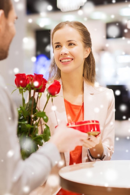 amor, romance, día de san valentín, concepto de pareja y gente - joven feliz con flores rojas dando regalos a una mujer sonriente en un café en un centro comercial con efecto de nieve