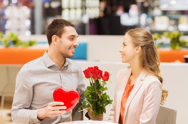amor, romance, día de san valentín, concepto de pareja y gente - joven feliz con flores rojas dando regalos a una mujer sonriente en un café en el centro comercial