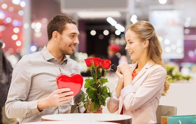 amor, romance, día de san valentín, concepto de pareja y gente - joven feliz con flores rojas dando regalos a una mujer sonriente en un café en el centro comercial