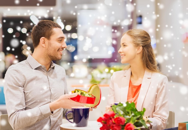 amor, romance, día de san valentín, concepto de pareja y gente - pareja joven feliz con flores rojas y caja de chocolate abierta en el centro comercial café con efecto de nieve